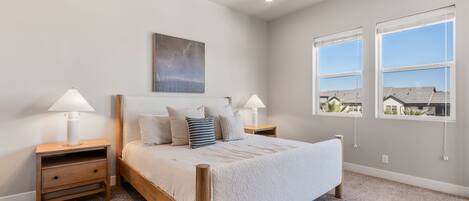 A bedroom with a wooden bed, two bedside tables with lamps, a large window with blinds, and a painting on the wall.