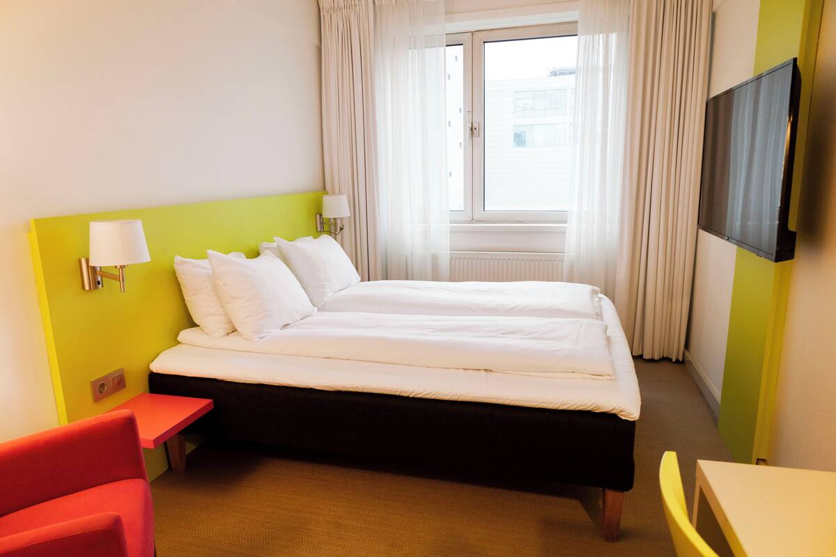A hotel room with a bed, a TV, a window with curtains, a red chair, and a small table.