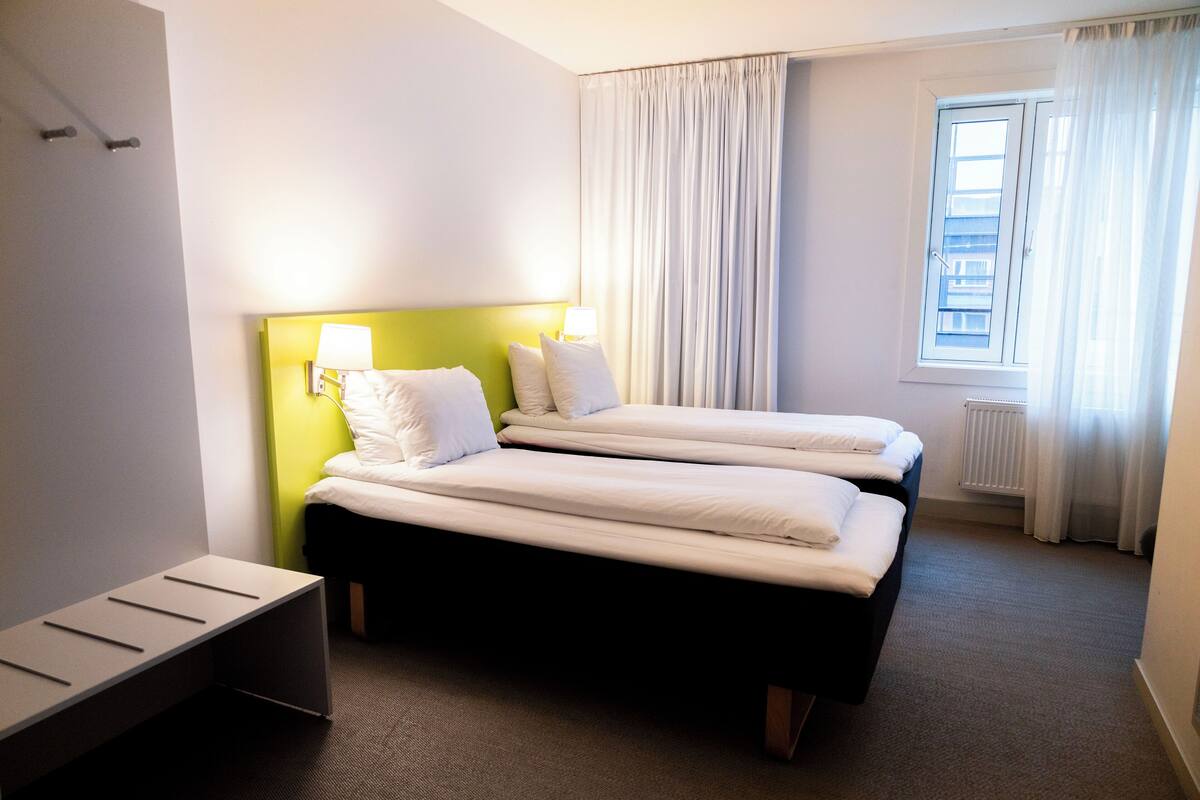 A hotel room with two beds, a bench, a window with curtains, and a radiator.