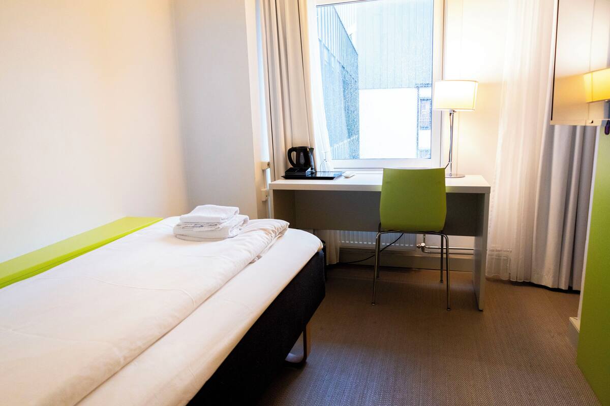 A hotel room with a bed, a desk, a chair, a lamp, and a window with curtains.