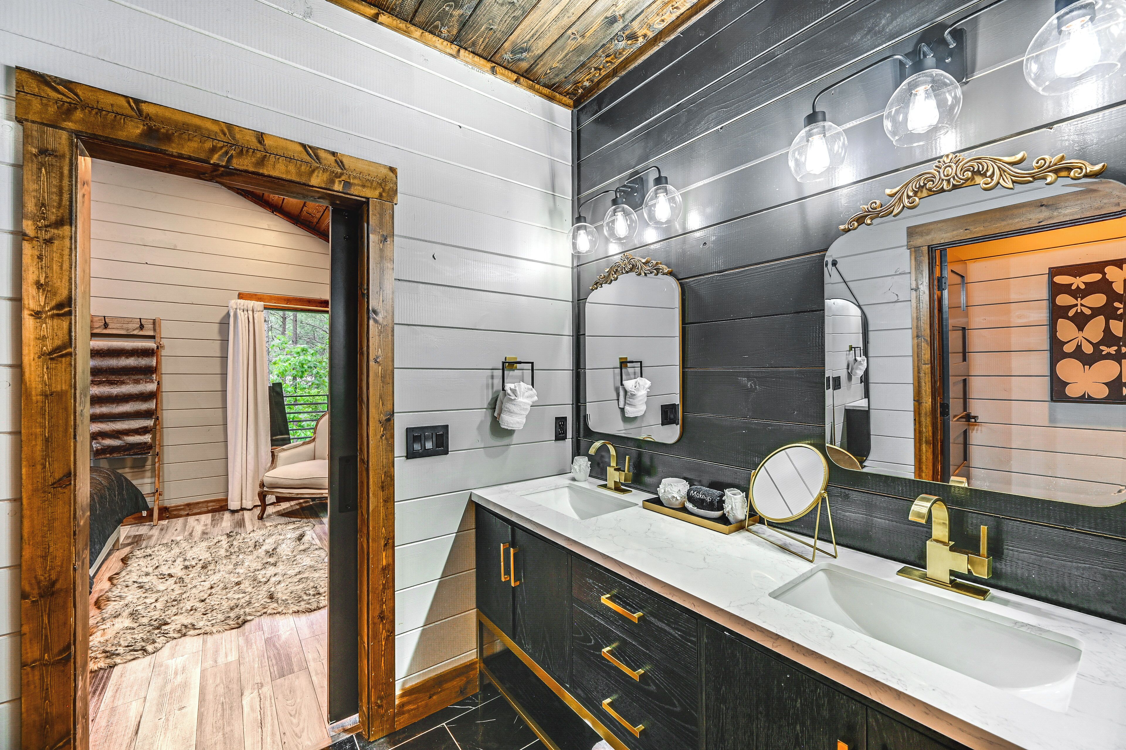 A bathroom with a large mirror, two sinks, and a wooden door leading to a room with a rug and a chair.