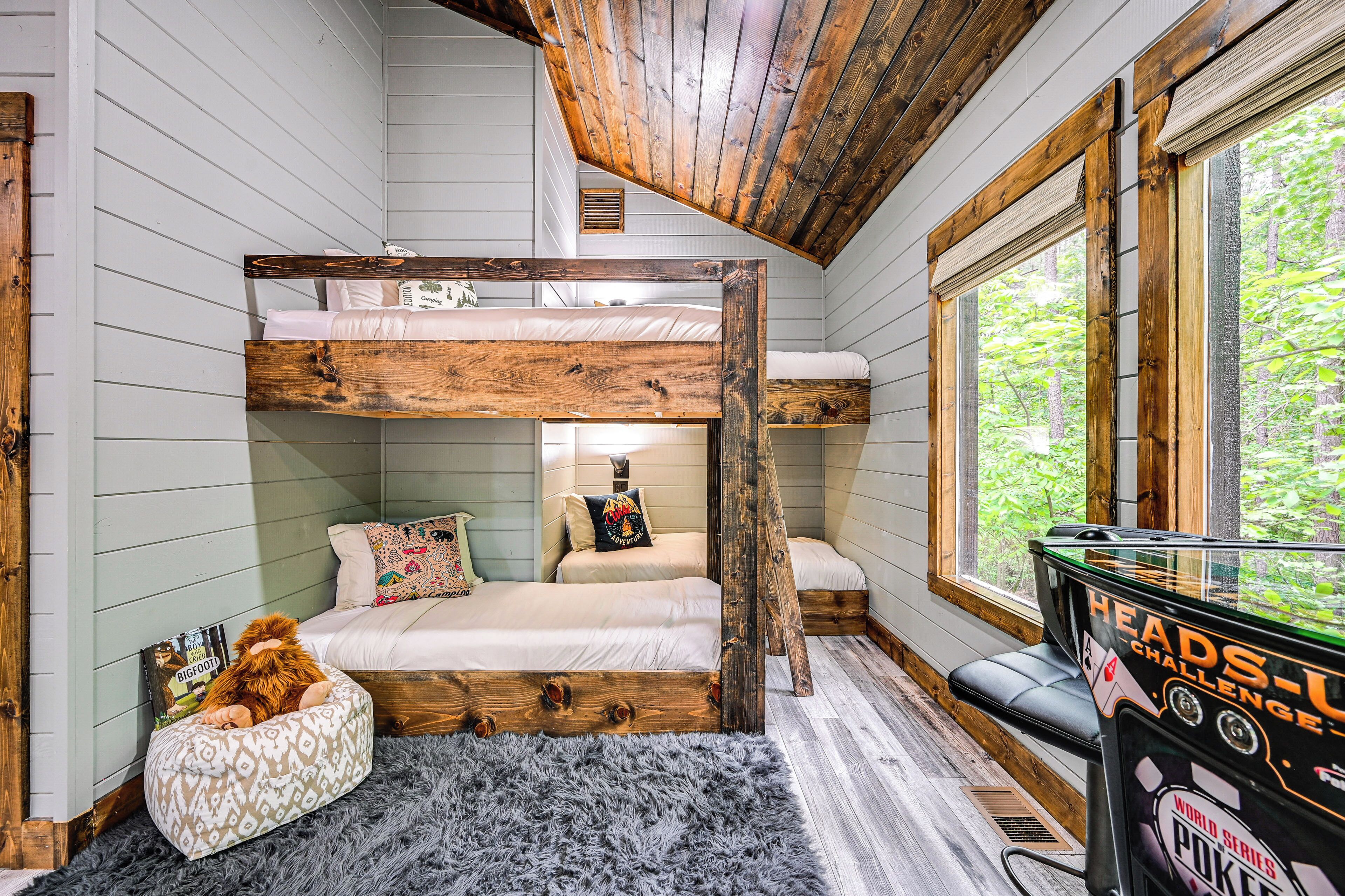 A bunk bed room with a wooden ceiling, a window with a view of trees, and a pool table.