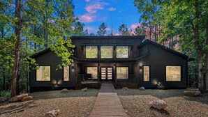 Une cabane en rondins moderne, dotée d’une terrasse en bois menant à l’entrée, entourée d’arbres et bordée d’un chemin de gravier.