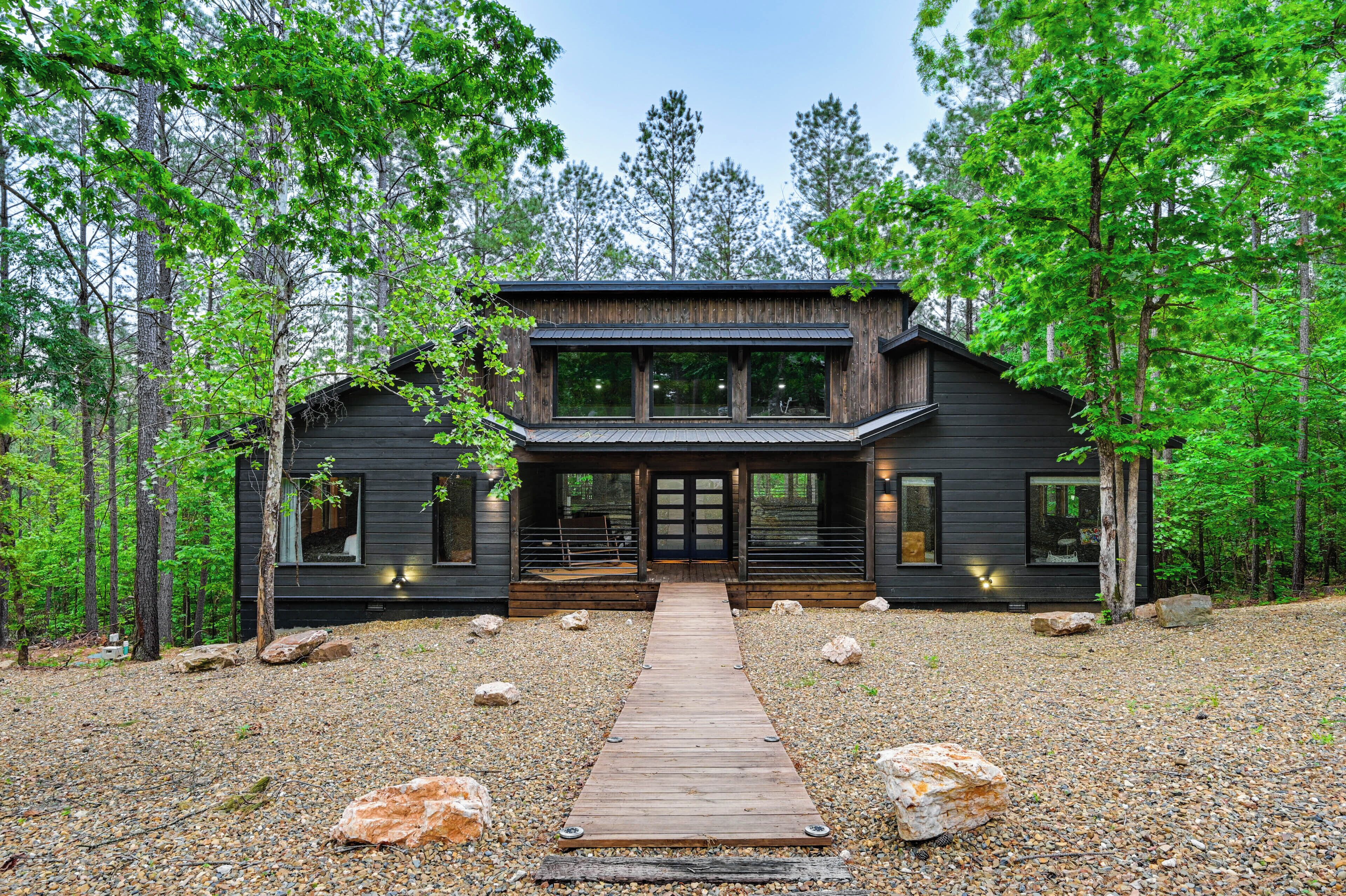 A modern log cabin with a wooden deck leading to the entrance, surrounded by a forest.
