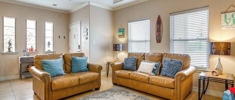 A living room with two leather sofas, a coffee table, and a rug. There are windows with blinds, a door, and wall decorations.