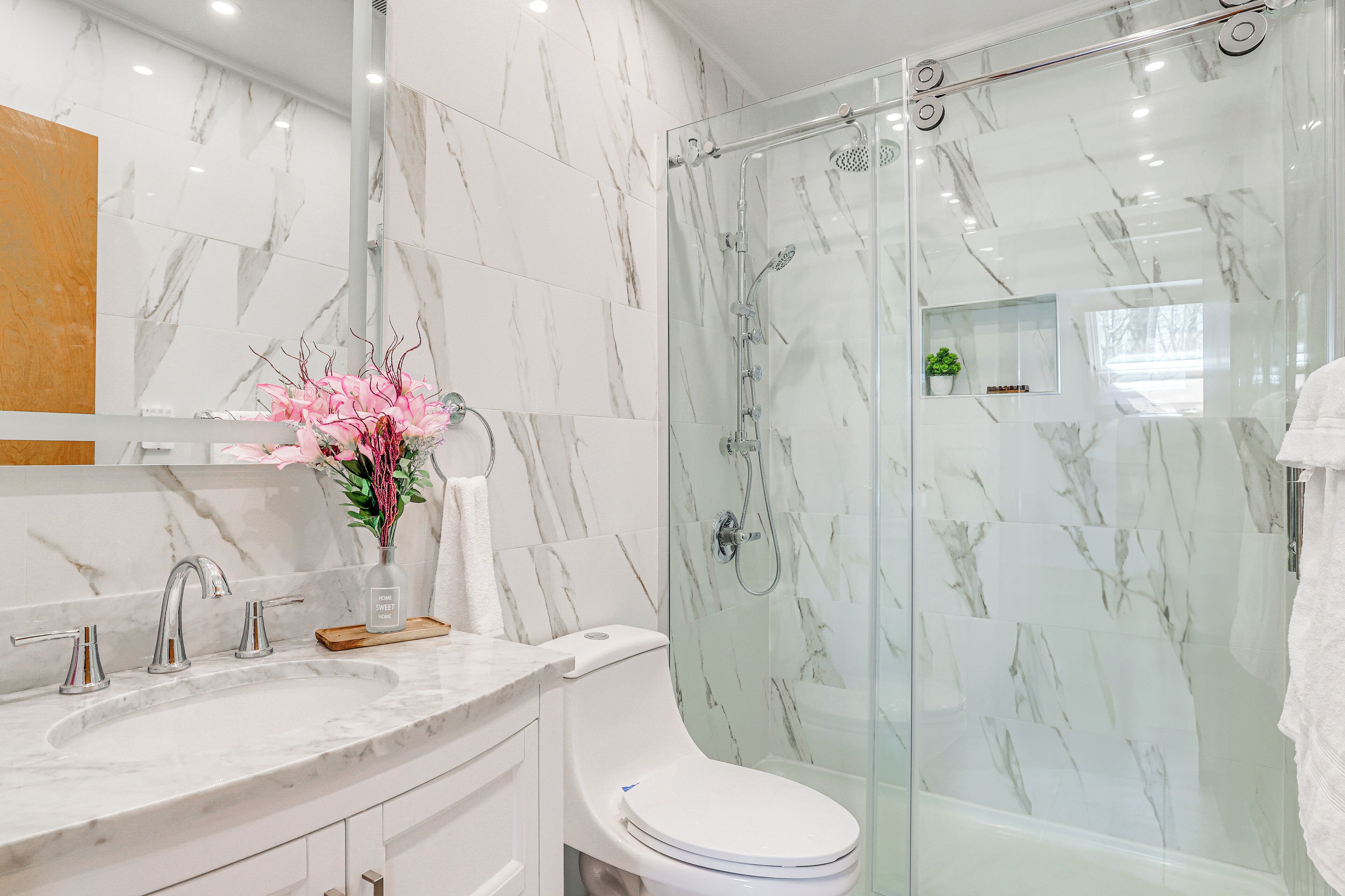 A bathroom with a marble vanity, a toilet, a glass shower enclosure, and a vase with flowers.