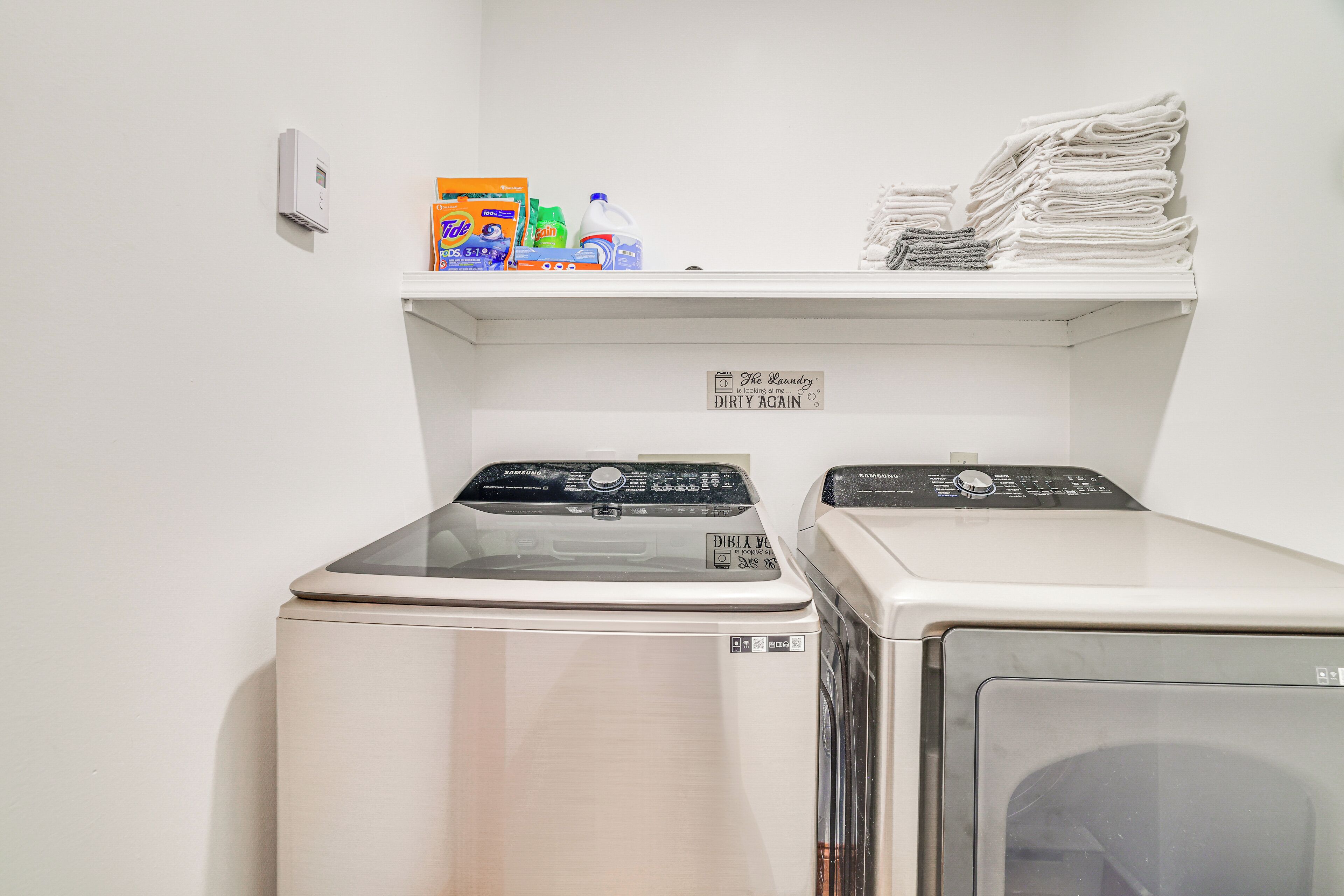 A laundry area with two washing machines, a shelf with detergent and folded towels, and a sign that says "The Laundry DIRTY AGAIN".
