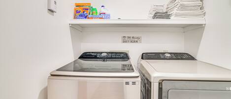 A laundry area with two washing machines, a shelf with detergent and folded towels, and a sign that says "The Laundry DIRTY AGAIN".