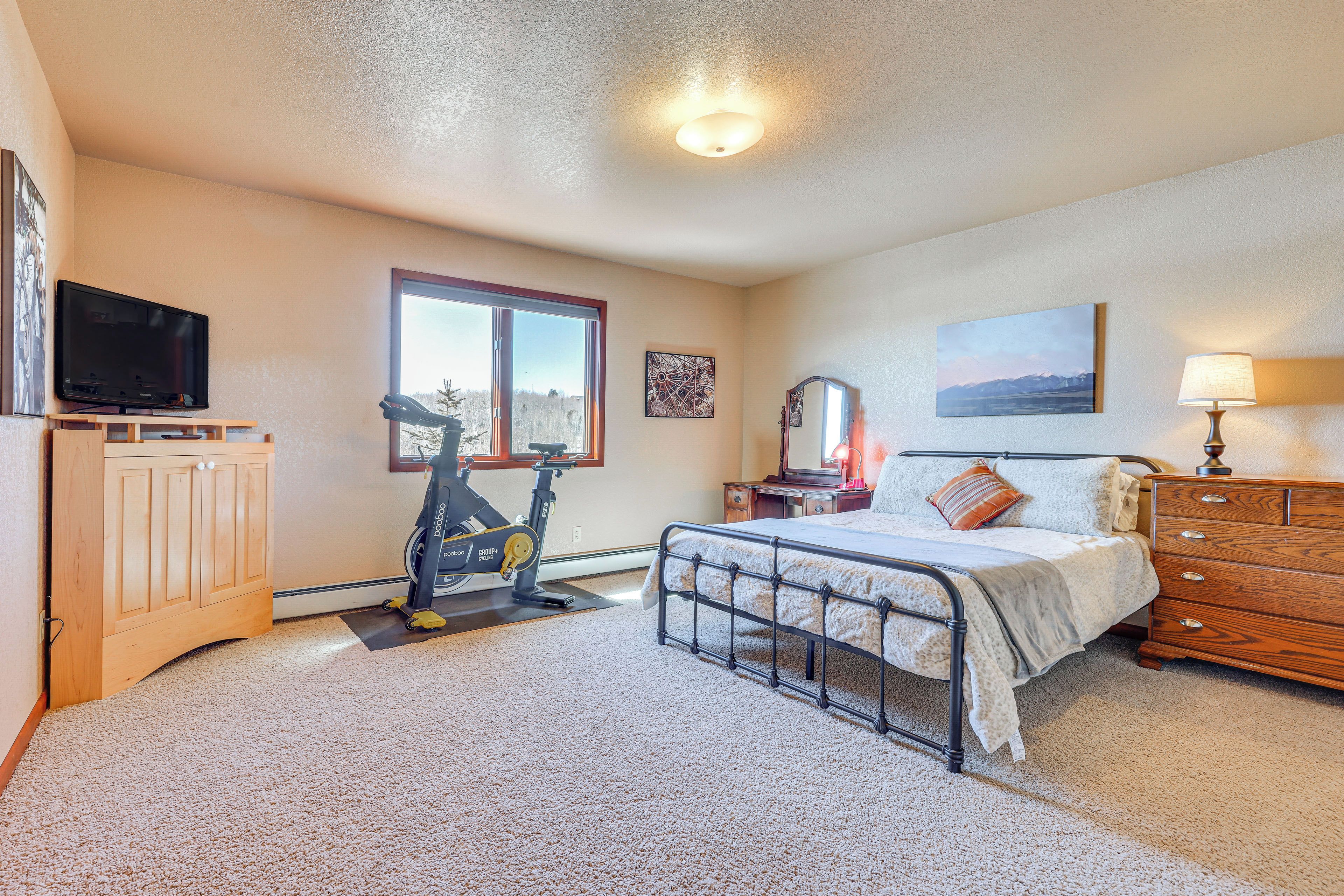 A bedroom with a bed, dresser, exercise bike, TV, and a window with a view.