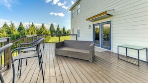 Une terrasse en bois avec un canapé et des fauteuils en osier, une table en verre et une maison aux murs recouverts de bardage blanc et à la porte bleue.