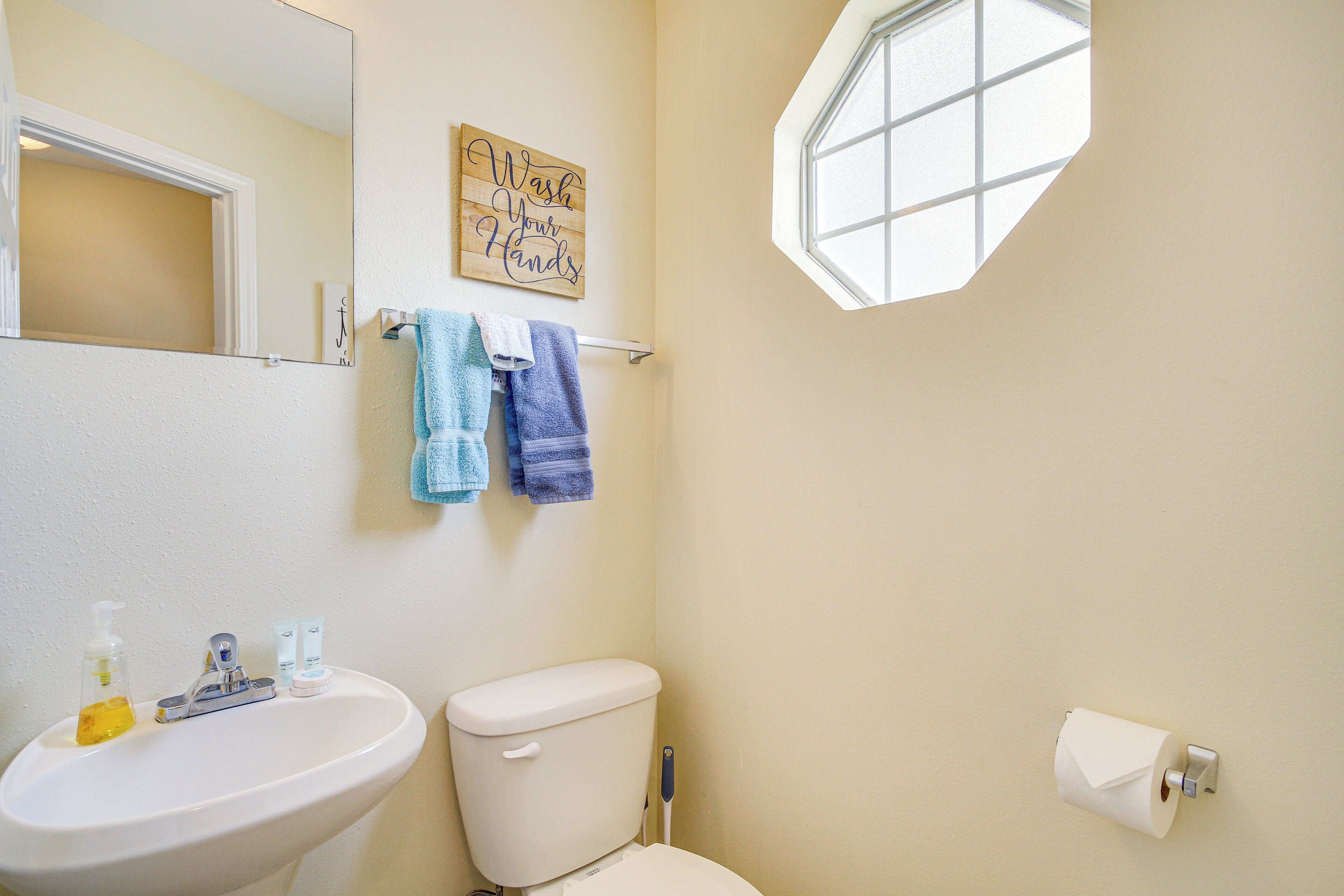 Une salle de bain avec un lavabo, des toilettes et une fenêtre. Il y a des serviettes accrochées au mur et une pancarte qui indique : « Lavez-vous les mains ».