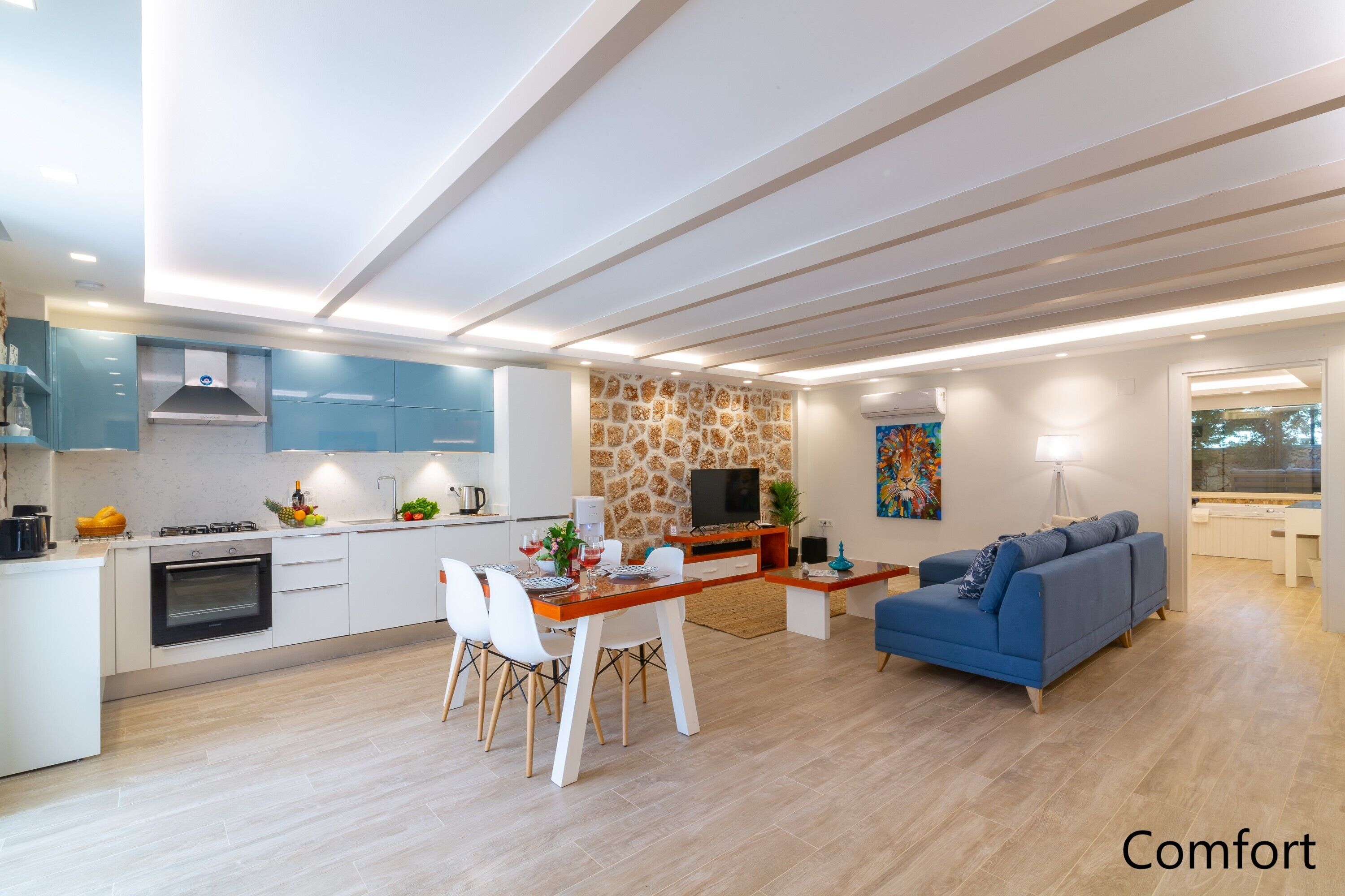 Una cuina moderna amb armaris blancs, una zona de menjador amb taula i cadires de fusta, un sofà blau i una paret d'accent de pedra.