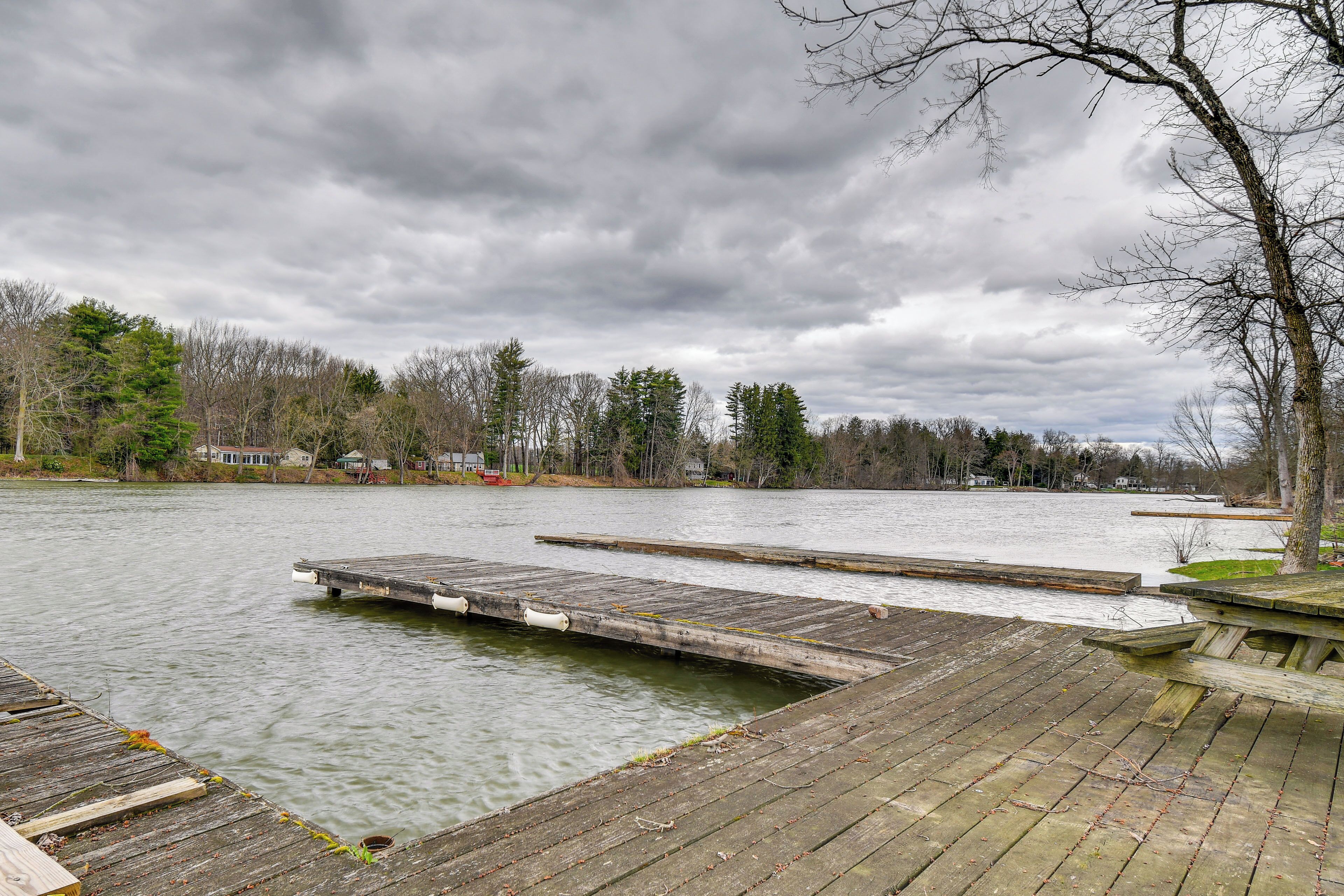 A wooden dock extending into a calm lake, surrounded by trees and houses in the background.