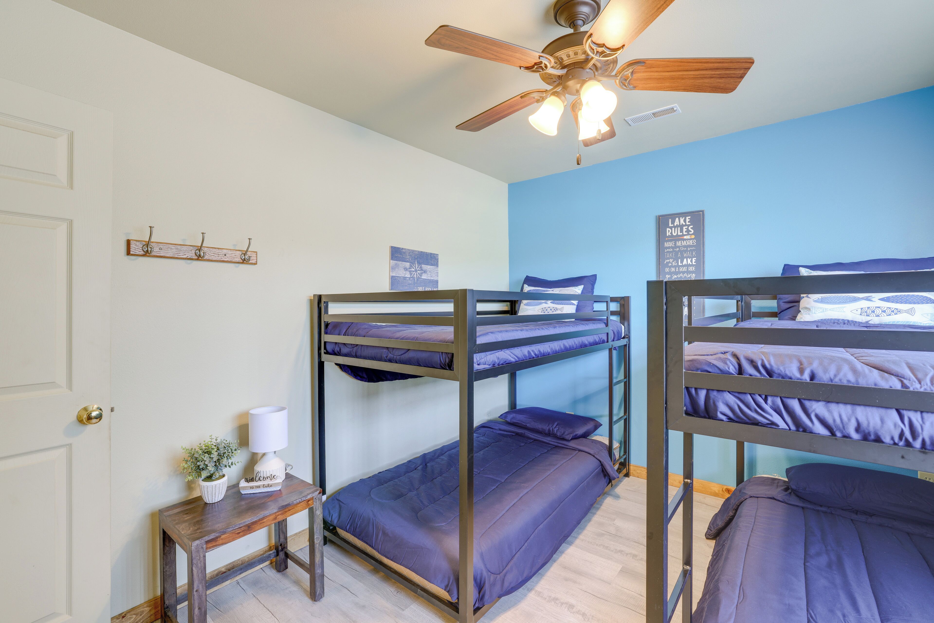 A dormitory room with bunk beds, a ceiling fan, a small table with a lamp, and a wall-mounted sign.