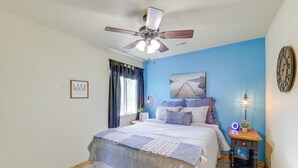 Un dormitorio con cama, ventilador de techo, mesita de noche con lámpara, reloj de pared y un cuadro enmarcado.