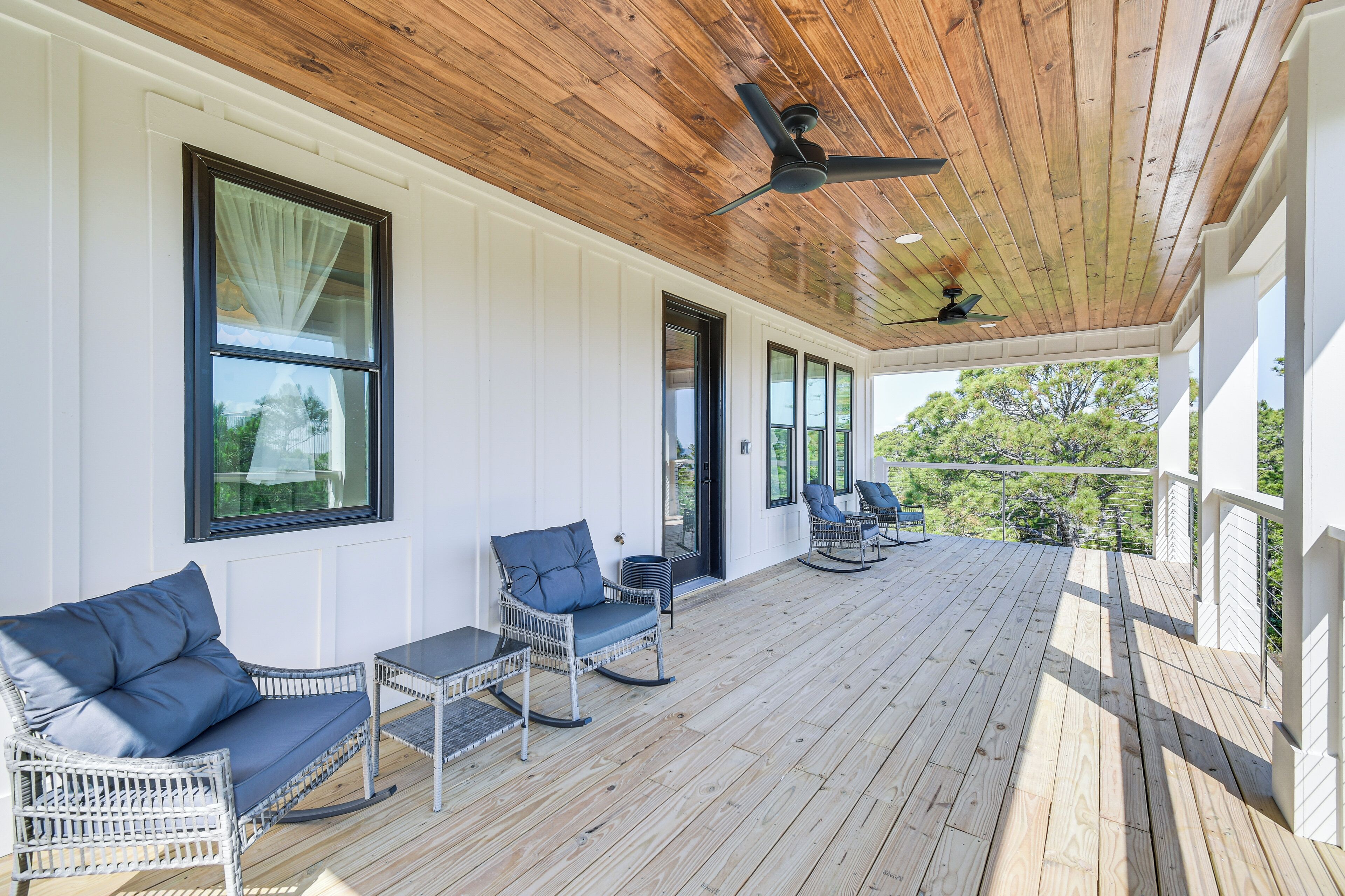 A covered porch with white walls, wooden ceiling, and ceiling fans. There are wicker chairs and a table with cushions.