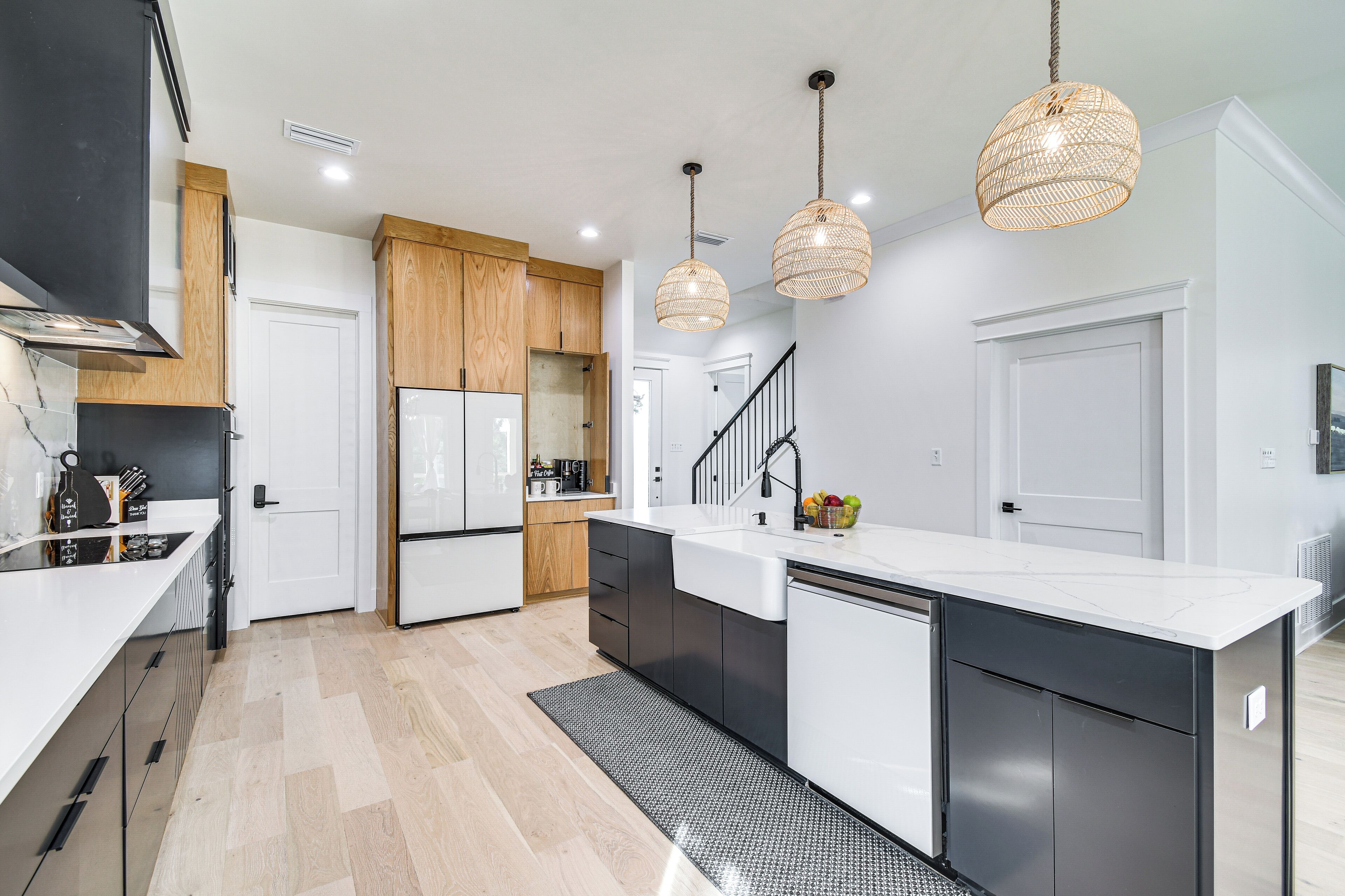 A modern kitchen with white countertops, black cabinets, and a wooden island. There are two hanging pendant lights and a staircase leading to another level.
