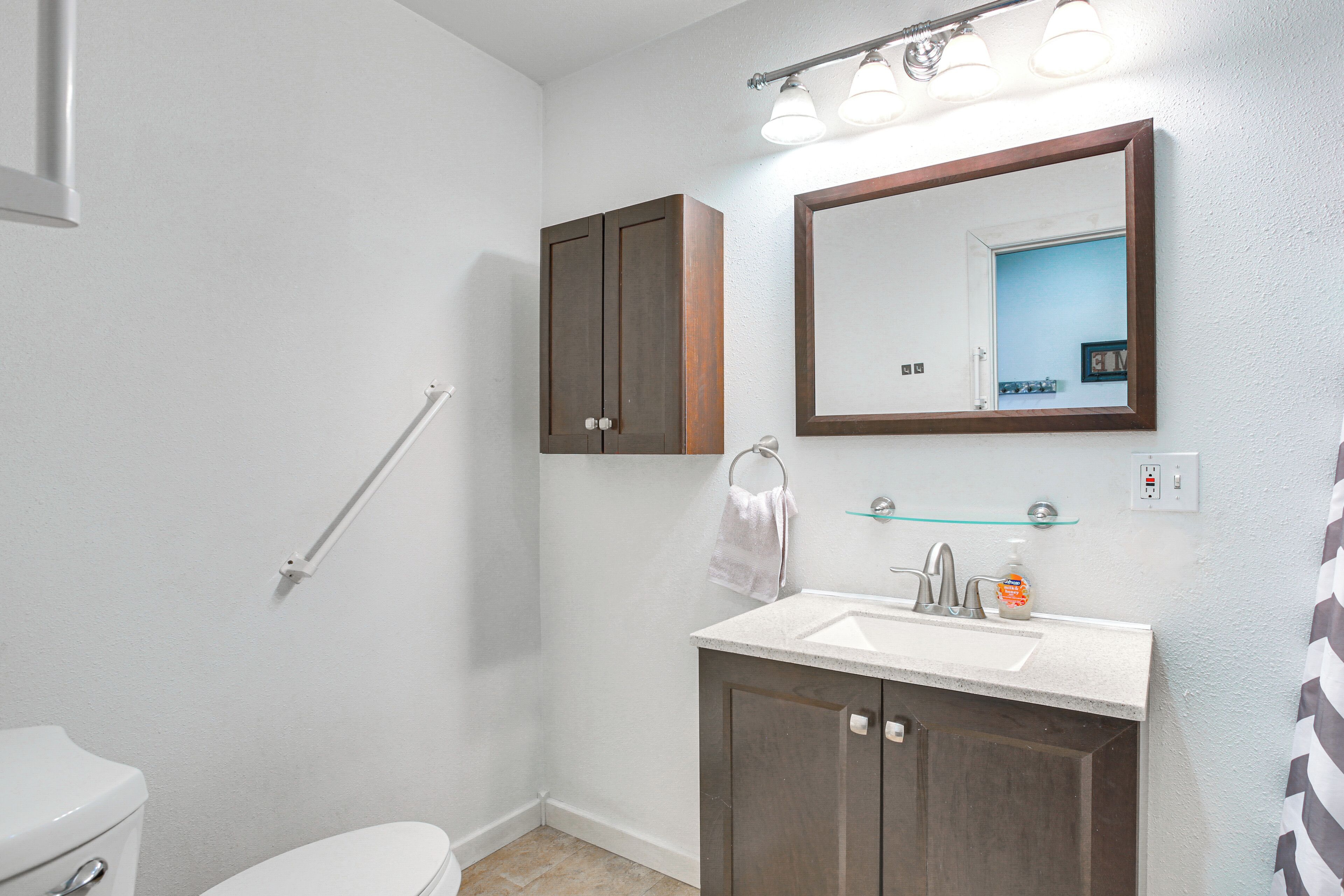 A bathroom with a white toilet, a dark wooden cabinet, a glass shelf with a bottle, and a mirror with a wooden frame.