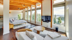 A spacious room with a large bed, a sofa, and a TV. There are multiple windows offering a view of trees and a lake. - Mid-century Escape: Lake Views, Steps to Cave Bay! (Worley)