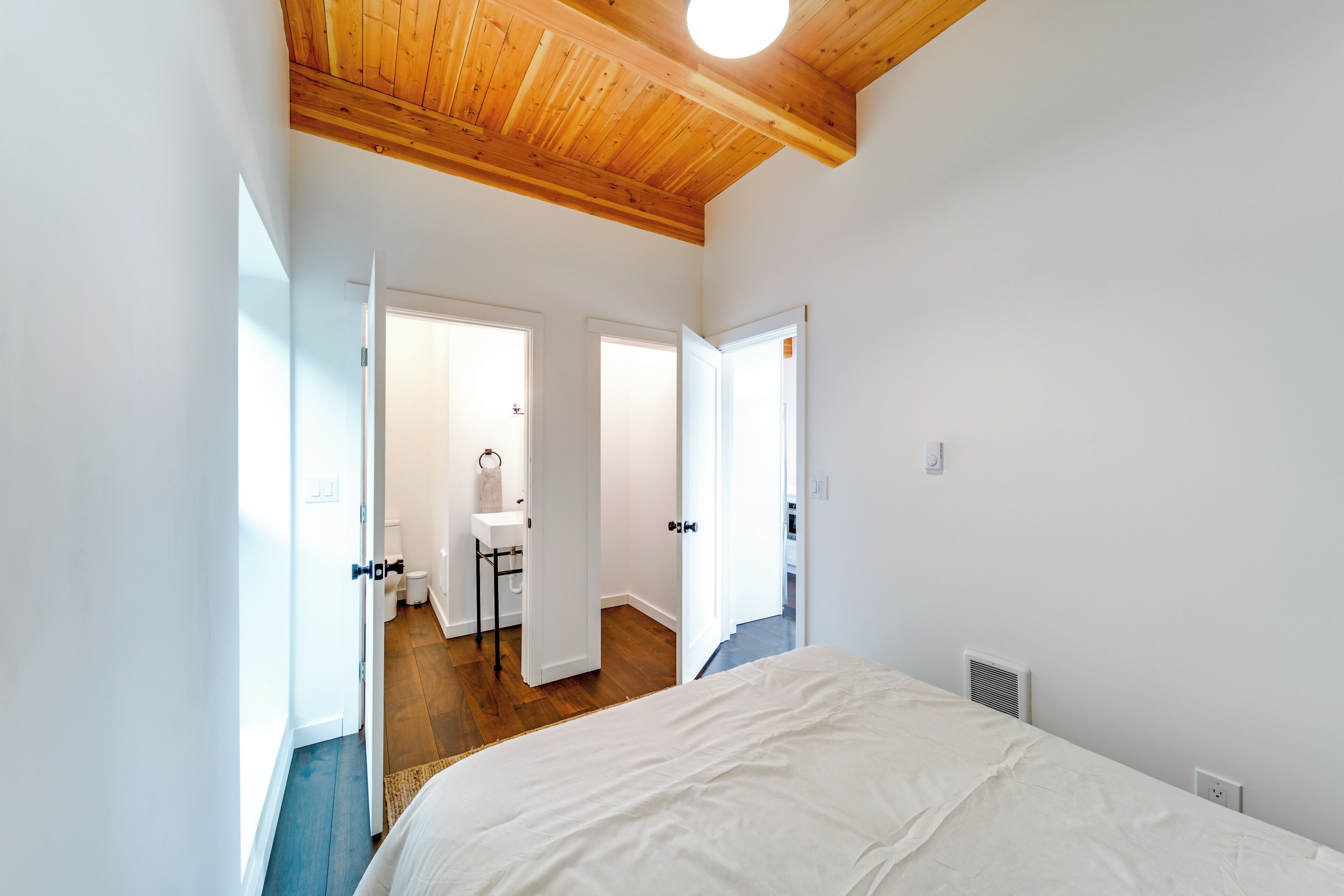 A bedroom with a wooden ceiling, white walls, and a bed with white bedding. There are two doors leading to other rooms.