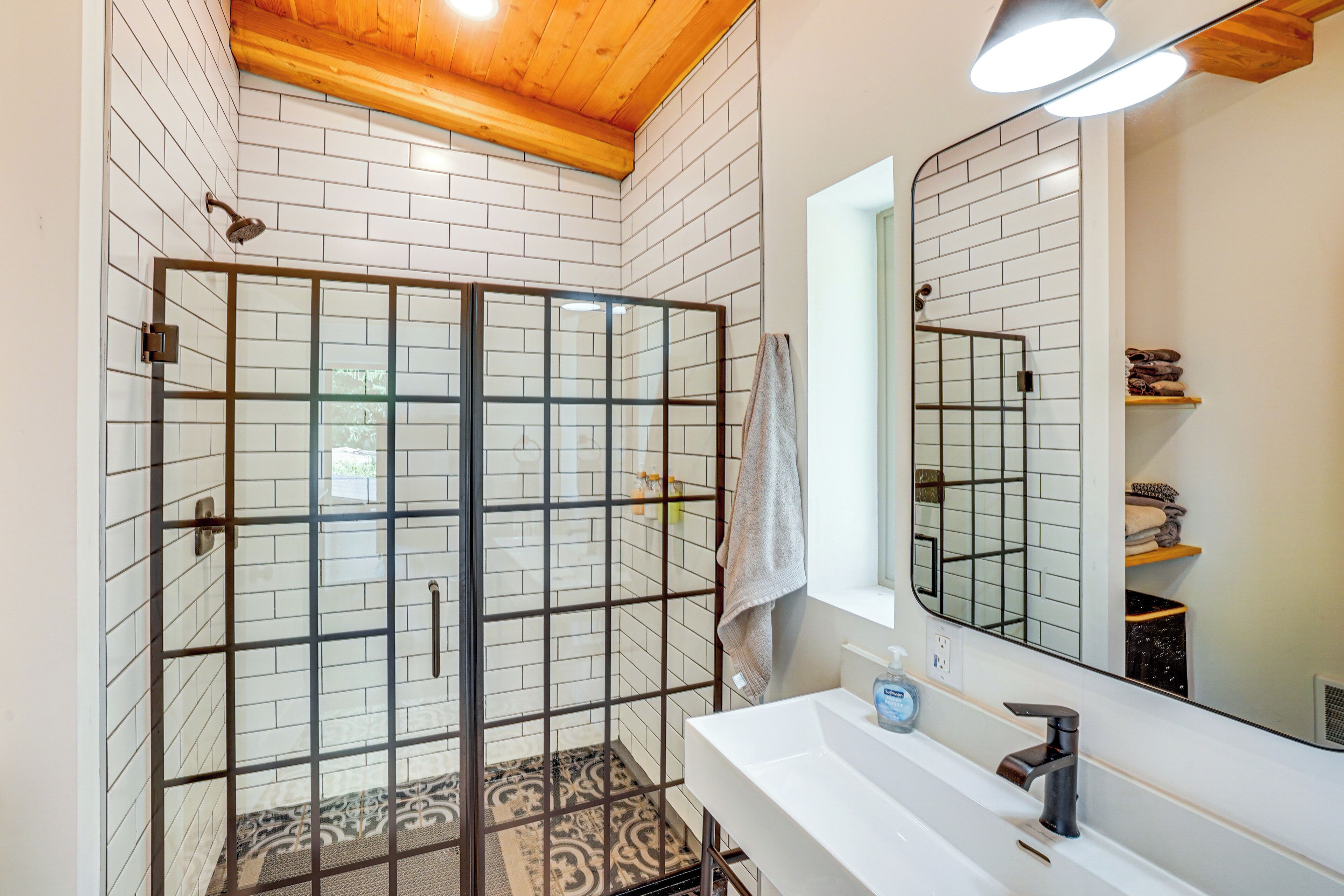 Un baño moderno con ducha de vidrio, azulejos blancos tipo metro, techo de madera, lavamanos blanco con grifo negro y espejo sobre el lavamanos.
