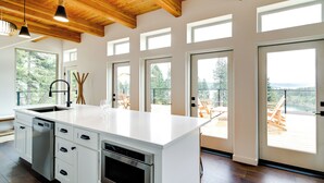 A modern kitchen with a large island, white cabinetry, and a wooden ceiling. There are multiple windows providing natural light and views of the outdoors. - Mid-century Escape: Lake Views, Steps to Cave Bay! (Worley)