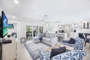 A modern living room with a large flat-screen TV, a grey sectional sofa, and a dining area with a kitchen in the background. - Waterfront Home | Table Tennis, Pool & Dock (Marco Island)