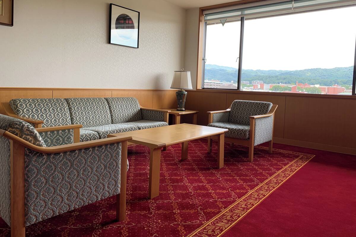 Une chambre d’hôtel avec un canapé, deux fauteuils, une petite table, une lampe et une grande fenêtre donnant sur un paysage urbain et des montagnes.