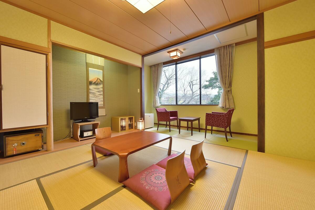 Une pièce de style japonais traditionnel, avec un sol recouvert de tatamis, une table basse, un téléviseur et une vue sur l’extérieur.