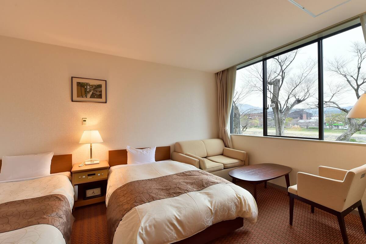 Une chambre d’hôtel avec deux lits, un canapé, une petite table et une vue sur les arbres et les bâtiments.