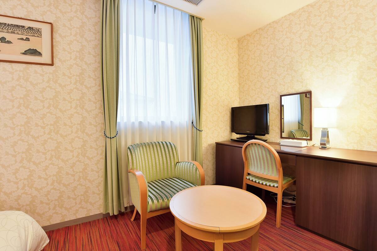 Une chambre d’hôtel comprenant un lit, un bureau avec une télévision, une chaise, une petite table et une fenêtre avec des rideaux.