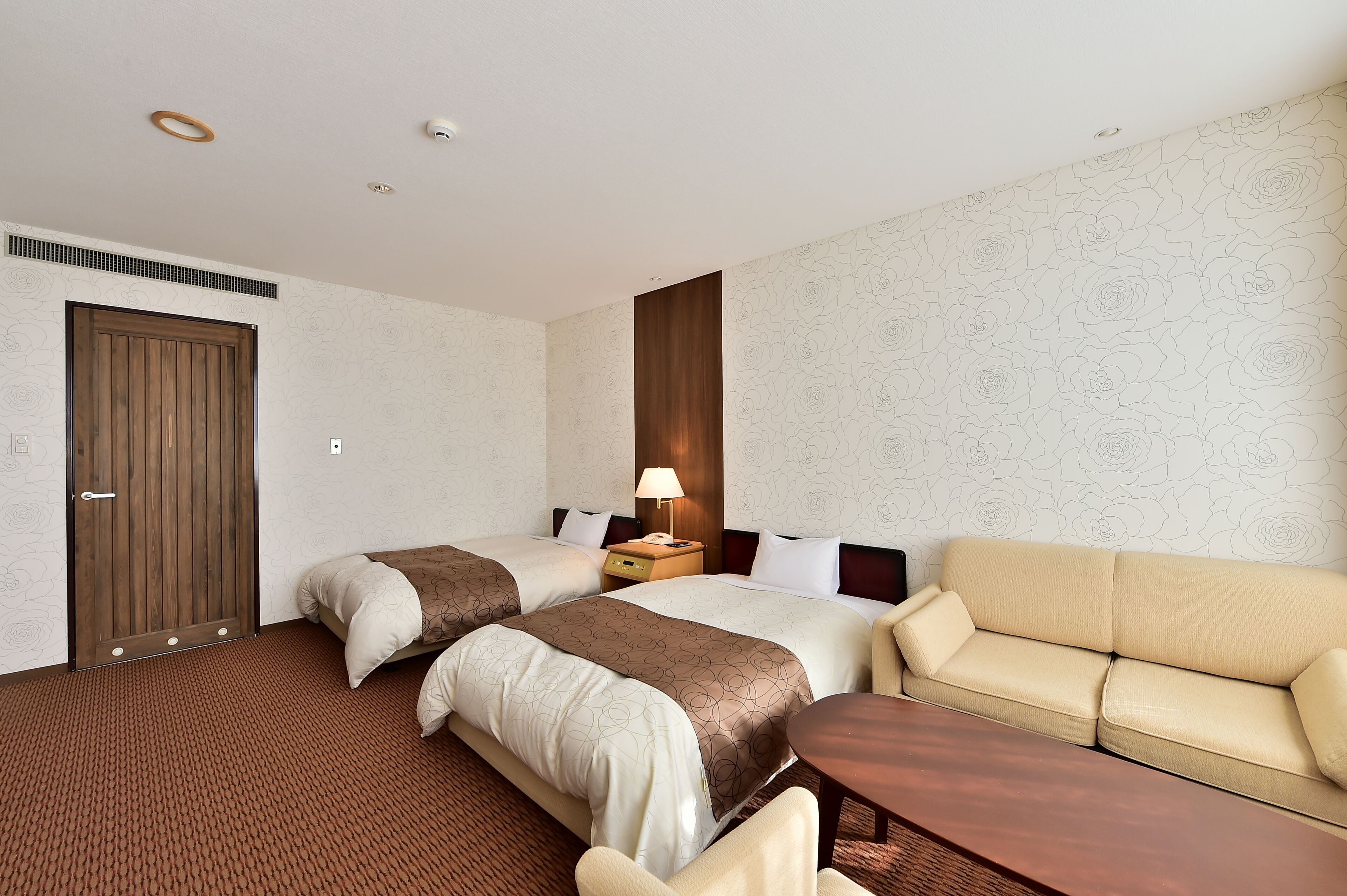A hotel room with two beds, a sofa, a wooden side table, and a lamp.