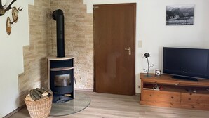 Un salon chaleureux avec un mur en pierre, un poêle à bois noir, une porte en bois, un téléviseur posé sur un meuble en bois et un panier de bois de chauffage.