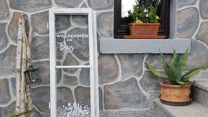 Un mur de pierre avec une pancarte de bienvenue, une petite fenêtre ornée d’une plante et un paillasson devant l’entrée.