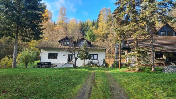 Une maison à deux étages, avec un toit recouvert de tuiles, entourée d’une cour verdoyante et d’arbres.