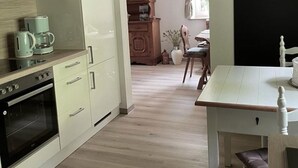 Une cuisine avec des meubles blancs, une machine à café et un four. On y trouve une table à manger avec des chaises et une fenêtre avec des rideaux.
