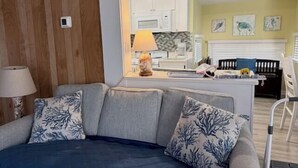 A modern living room with a grey sofa, blue patterned cushions, a lamp, and a view into the kitchen area. - 1050 Ocean Lakes Pet Friendly Golf Cart⛱️🏝️🩱🌊👙☀️🐚 (Myrtle Beach)