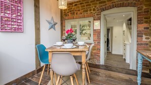 Un coin repas avec une table en bois, des chaises bleues et grises, un objet décoratif rose accroché au mur et un mur en briques avec une porte en verre.