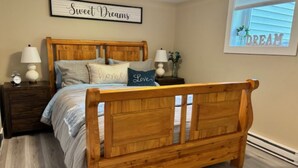 Une chambre à coucher avec un lit en bois, deux tables de chevet, un panneau décoratif fixé au mur et une fenêtre avec des stores.