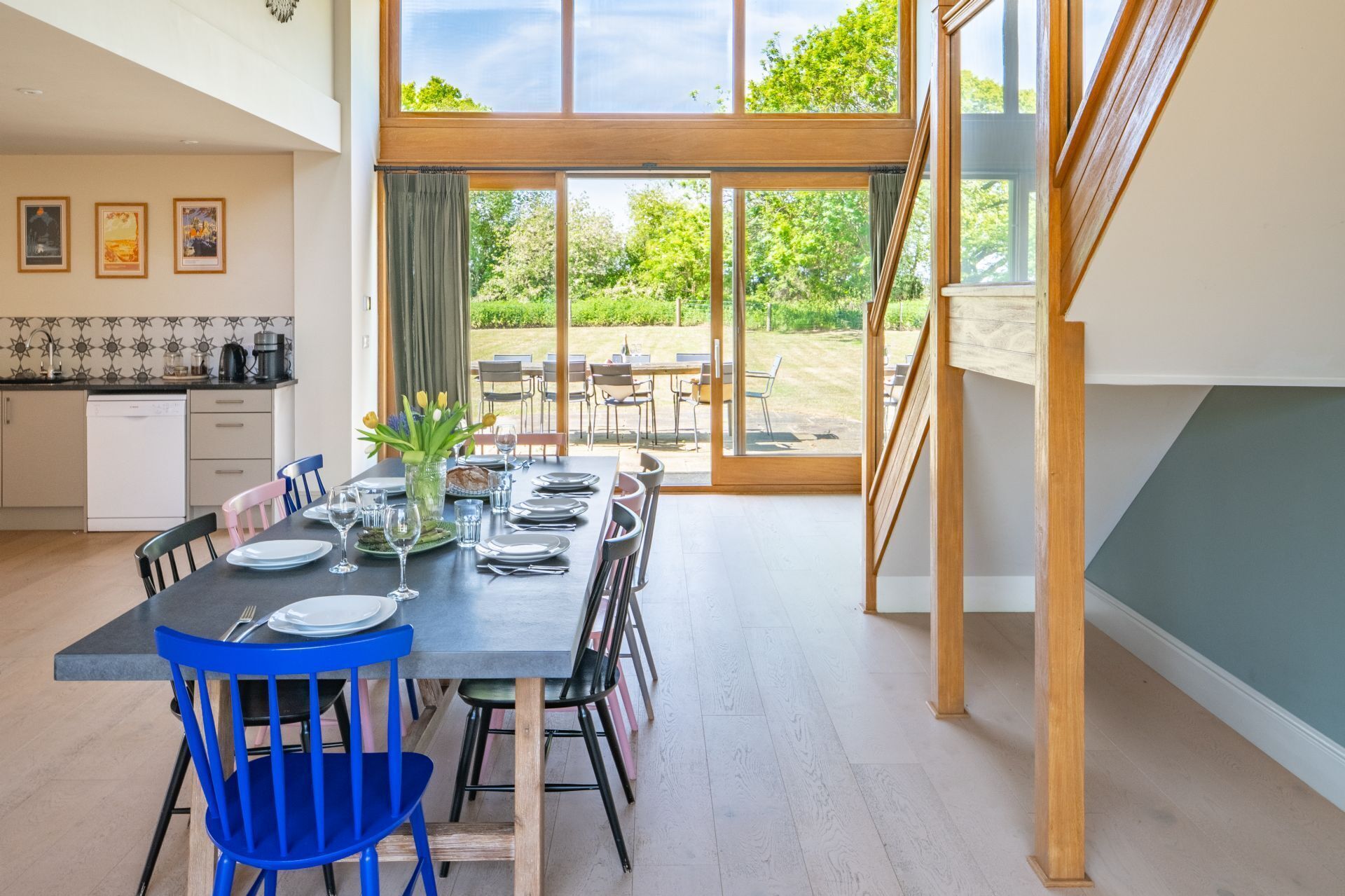 A dining area with a table set for six, a kitchen in the background, and a view of the outdoors.
