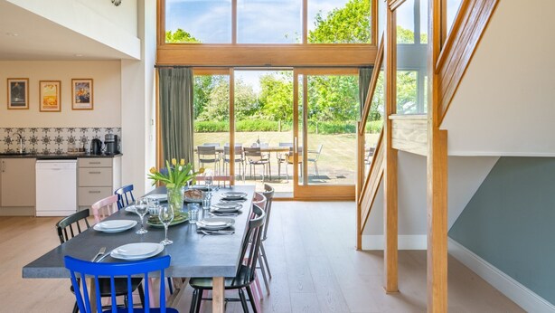 Un coin repas avec une table dressée pour six personnes, une cuisine en arrière-plan et une vue sur l’extérieur.