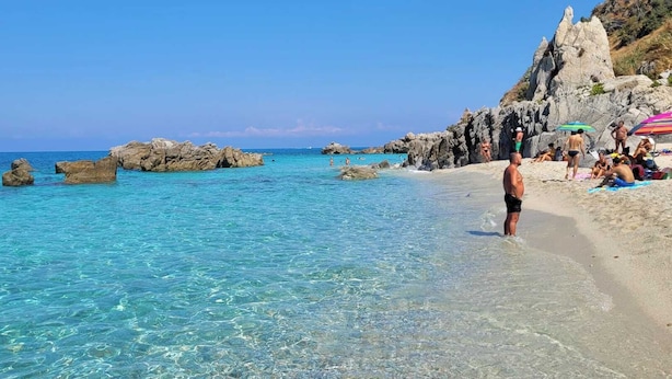 Una spiaggia con acque turchesi cristalline, formazioni rocciose e persone che prendono il sole.