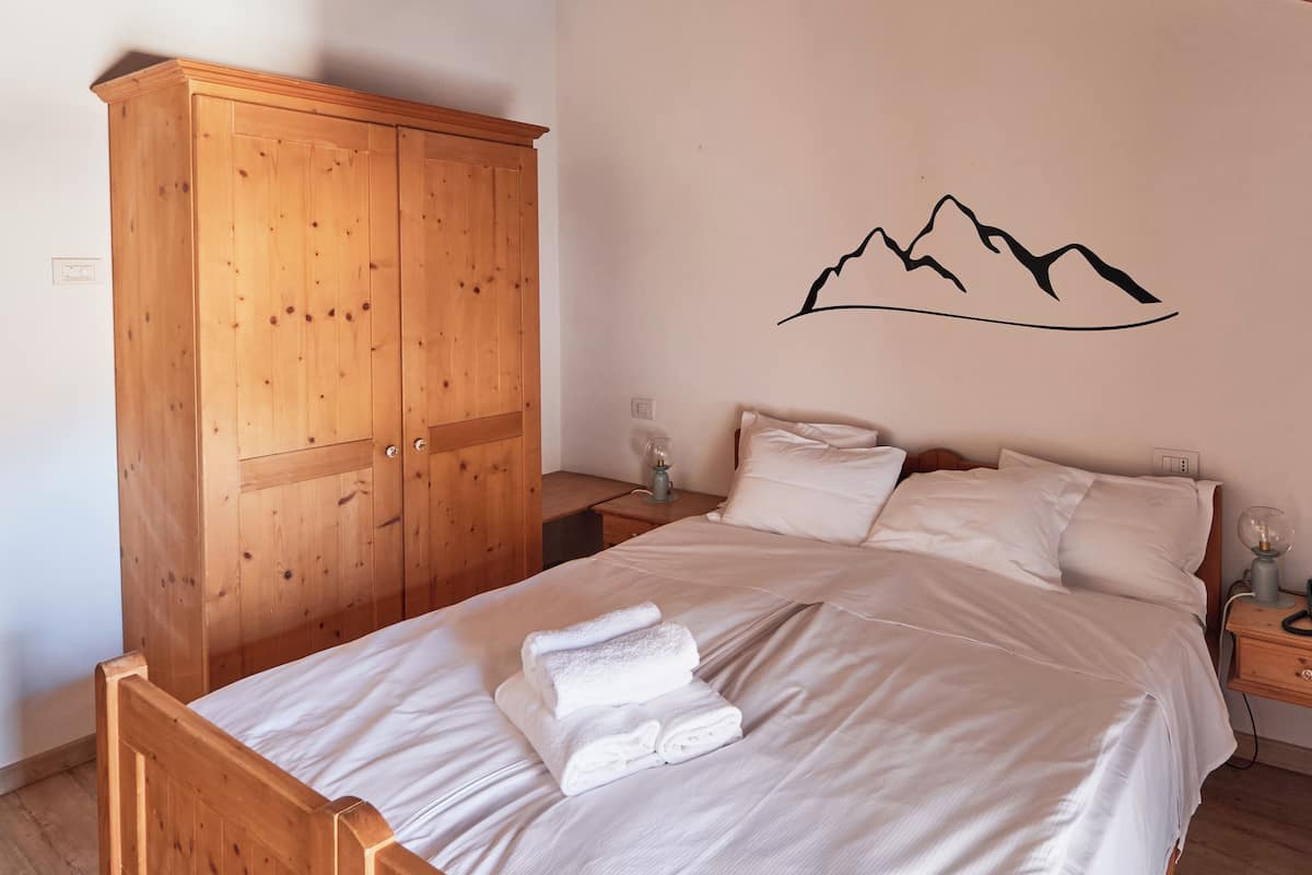 A bedroom with a wooden wardrobe, a bed with white linens, and a wall decal of mountains.
