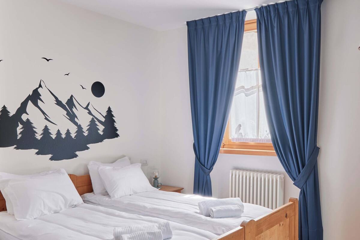 A bedroom with a wooden bed, white bedding, a mountain-themed wall decal, a window with blue curtains, and a radiator.