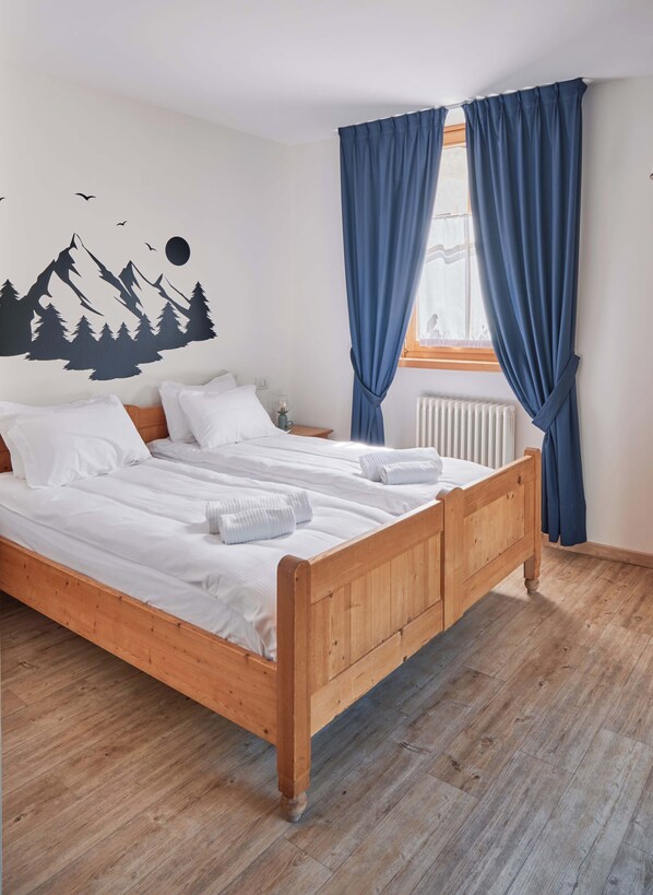 A bedroom with a wooden bed, white bedding, a mountain-themed wall decal, a window with blue curtains, and a radiator.
