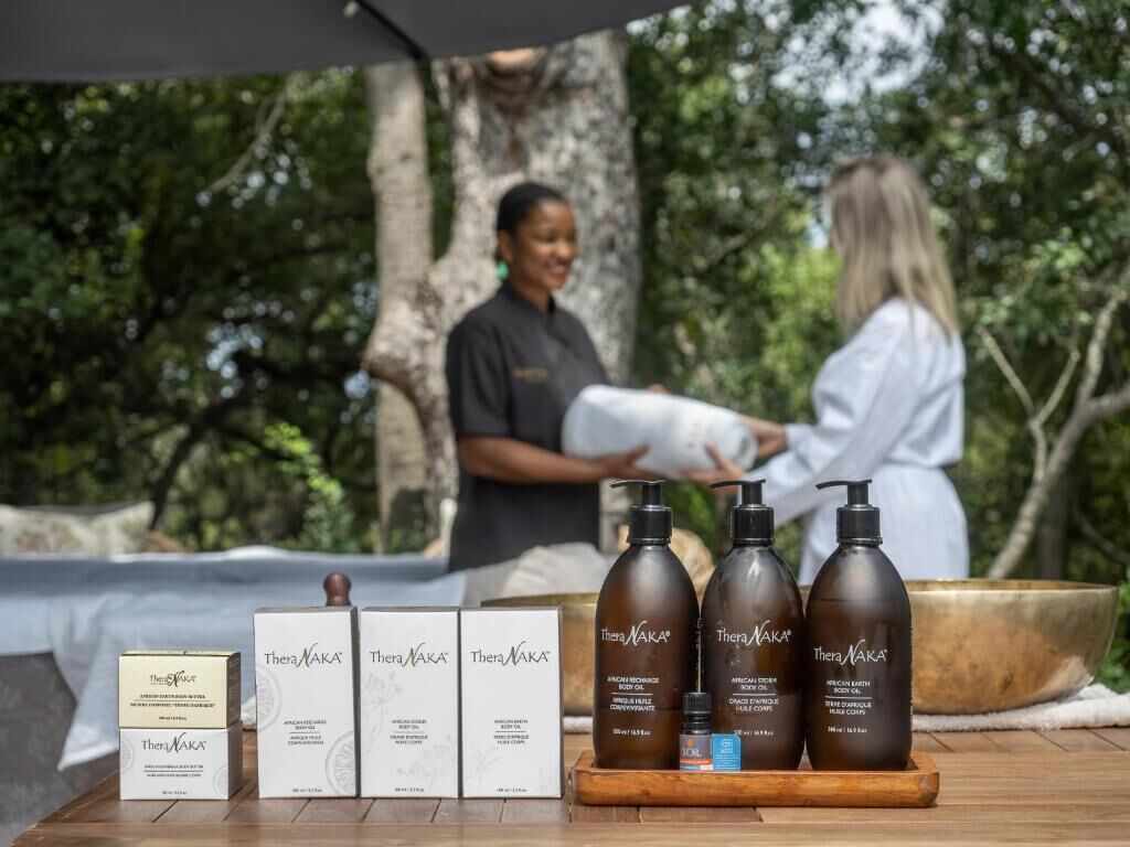 Un ambiente de spa con una mesa de madera que exhibe productos Theravaka, una camilla de masaje y una persona vestida con una bata blanca.