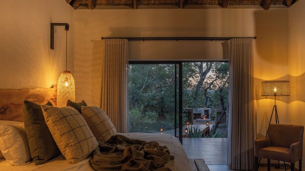 Quarto com cama grande, uma cadeira, um abajur e vista para o exterior.