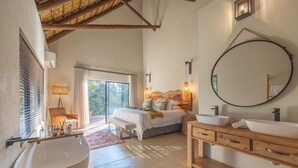 Quarto moderno com uma banheira grande, uma toucador de madeira, um espelho redondo e uma cama com vista para o exterior.
