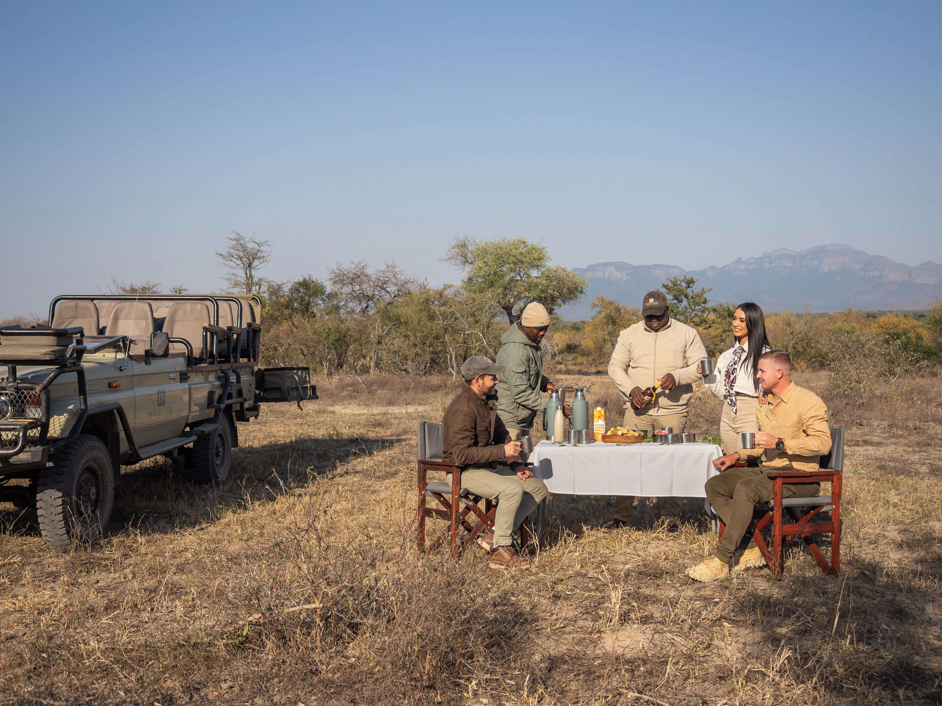 A group of people seated around a table in an outdoor setting with a vehicle nearby.