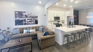 A modern living room with a sectional sofa, a coffee table, and a kitchen area with white cabinets, a microwave, and a large island with bar stools.